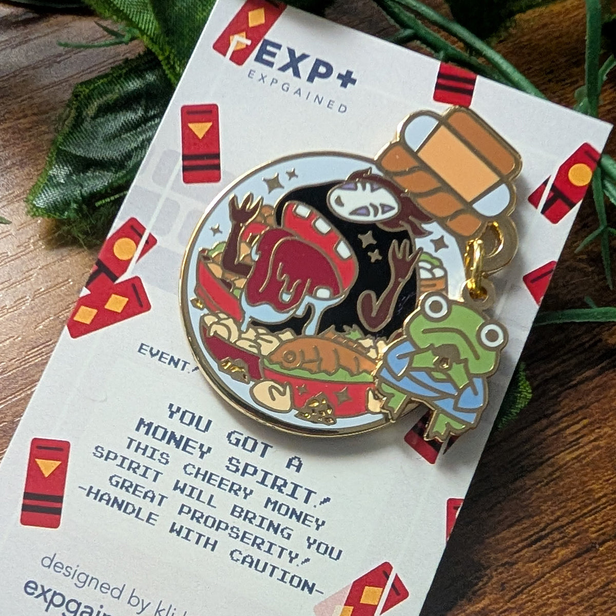 An enamel pin featuring a no face character eating and a green frog in a blue suit, attached to a gold ring and displayed on its white EXP+ Expgained backing card over a wooden surface with green leaves.