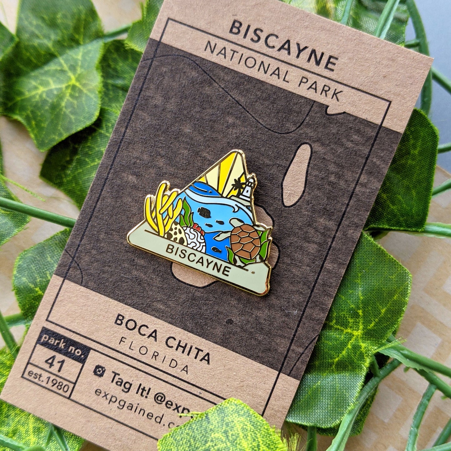 Triangle Biscayne national park enamel pin featuring a view from the beehive hike on a brown business card size backing card with a topo map of the underwater life and the Boca Chita lighthouse in the distance.