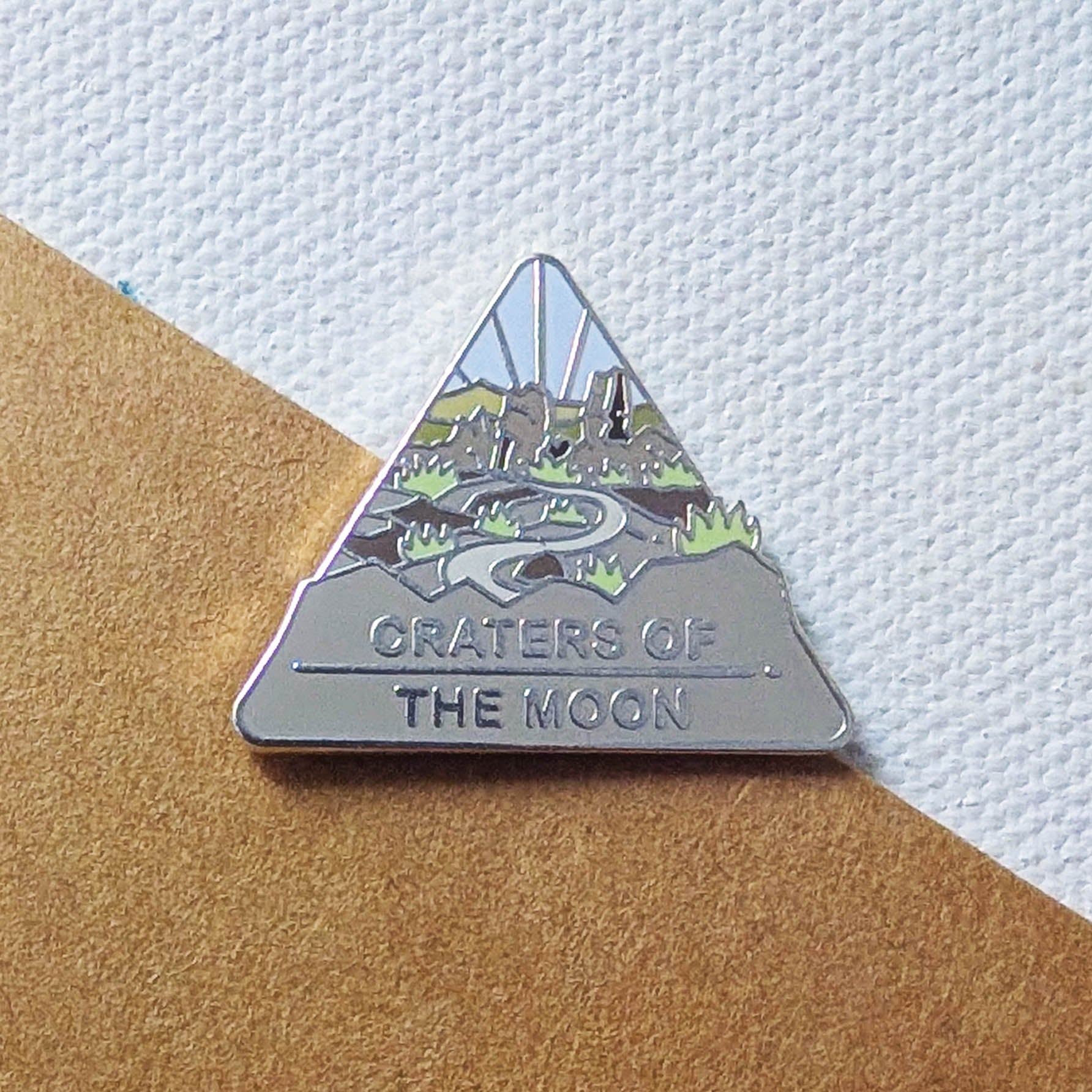 Triangular enamel pin with 'Craters of the Moon' design on a textured surface