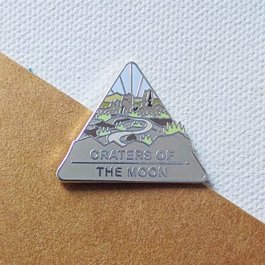 Triangular enamel pin with 'Craters of the Moon' design on a textured surface