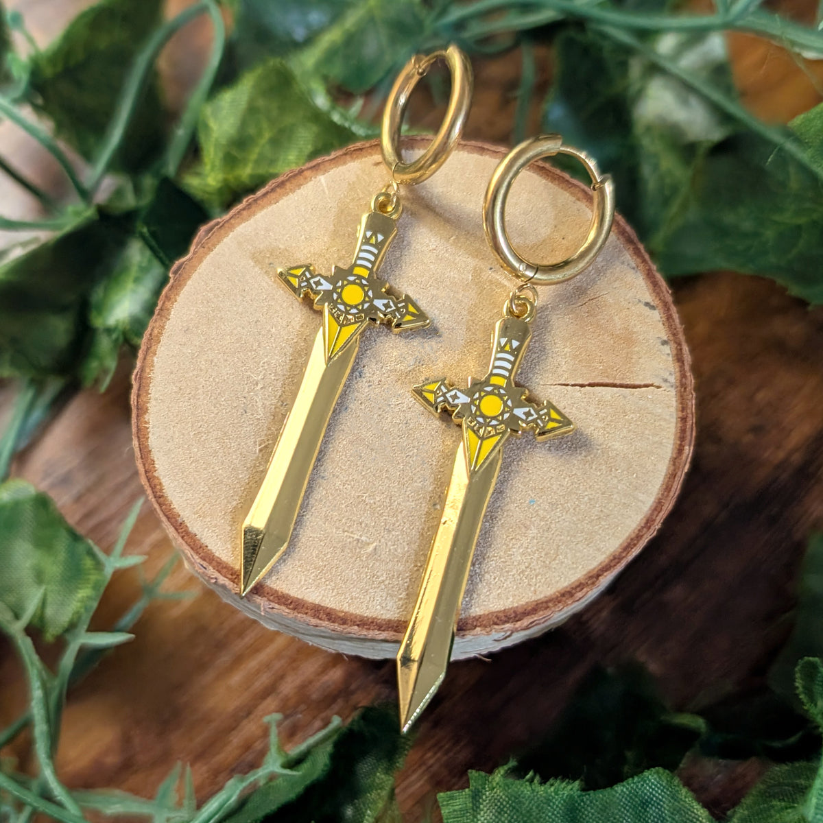 A pair of gold-tone dangle earrings shaped like miniature fantasy swords with circular hoop tops, displayed on a round wood slice surrounded by green leaves.