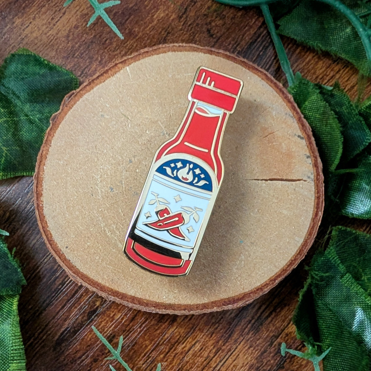 A small enamel pin shaped like a hot sauce bottle, placed on a round wood slice with green leaves around it on a wooden surface.
