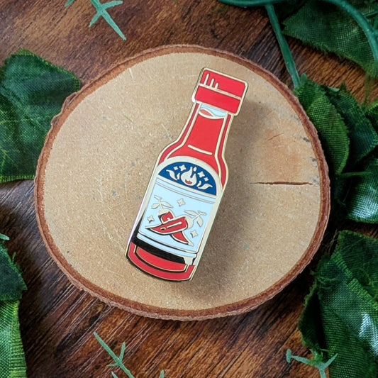 A small enamel pin shaped like a hot sauce bottle, placed on a round wood slice with green leaves around it on a wooden surface.