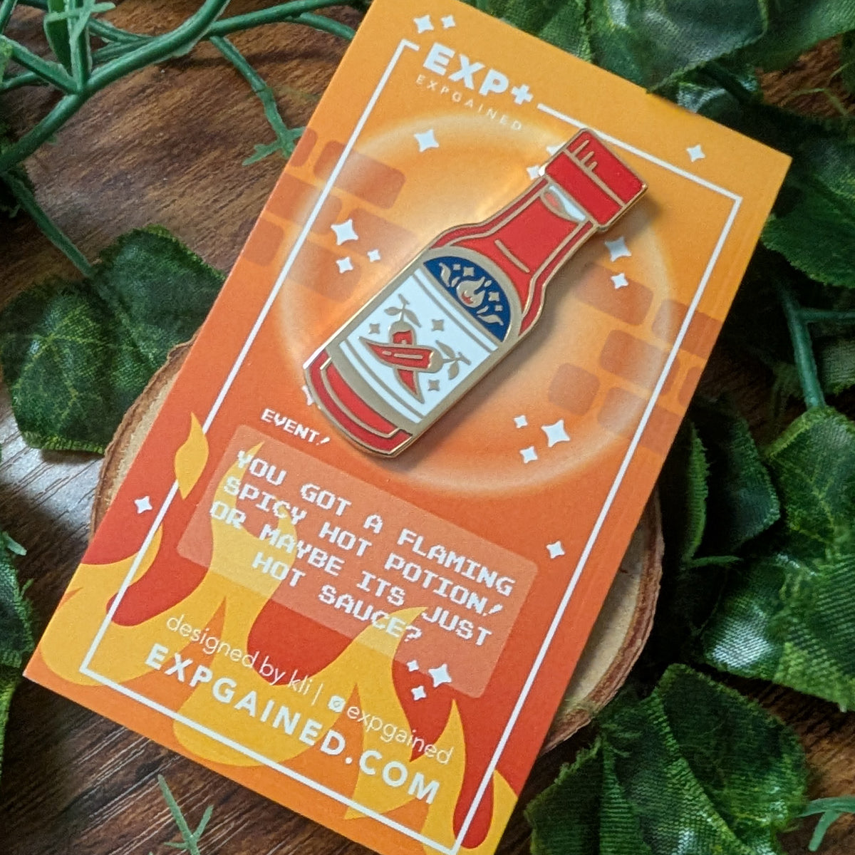 An enamel pin shaped like a hot sauce bottle is displayed on its orange backing card, which features text and flames, set against a background of green leaves and a wooden surface.