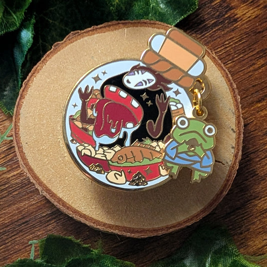 A circular enamel pin featuring a whimsical scene with a red mushroom, a fish, and a green frog character, attached to a gold-tone ring and displayed on a round wood slice with green leaves in the background.
