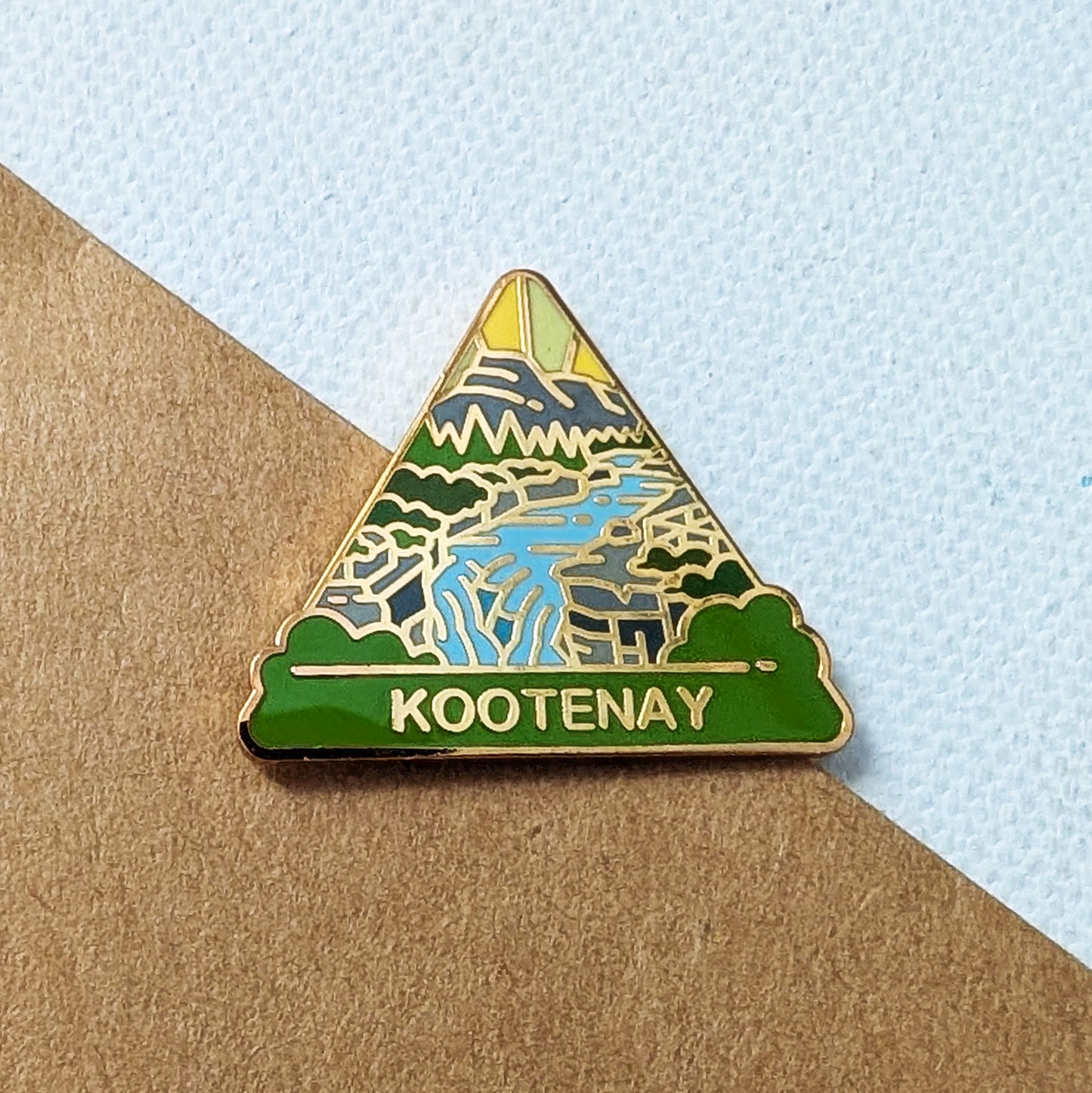 A triangular enamel pin featuring a stylized landscape with mountains, trees, a river, and the word 'KOOTENAY' on a green base, photographed on a brown paper surface against a pale blue textured background.
