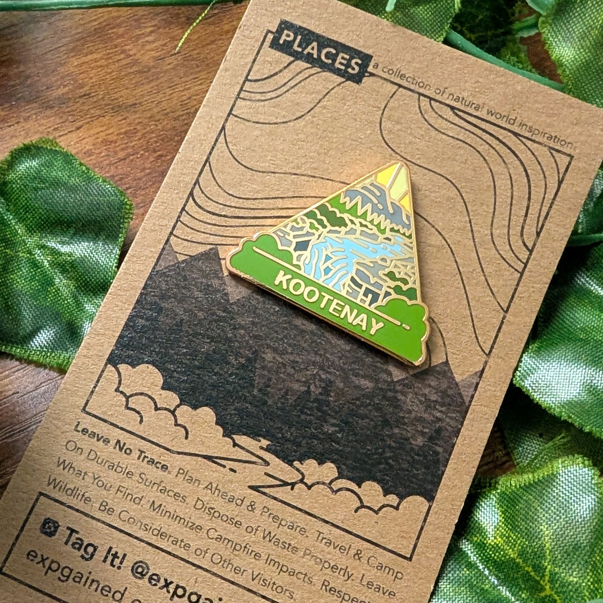 A triangular enamel pin featuring a mountain landscape with the text 'KOOTENAY' on a kraft paper backing card, styled with green leaves on a wooden surface.