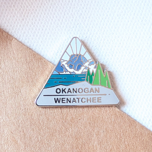 Triangular enamel pin with mountain and lake design on a textured surface.