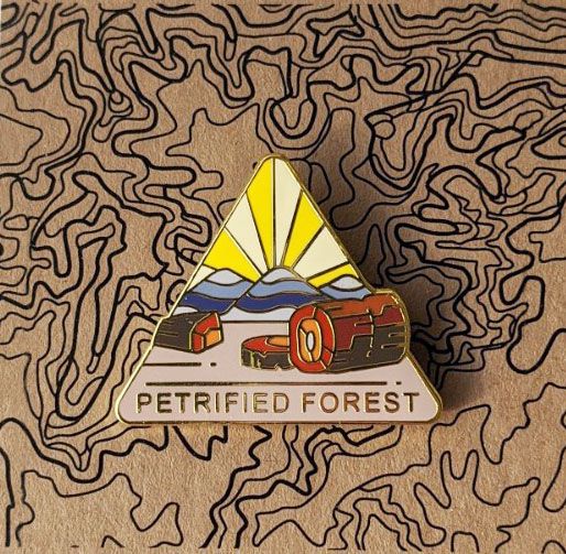 Triangle Petrified Forest national park enamel pin featuring a view of petrified wood and blue hills in the background.