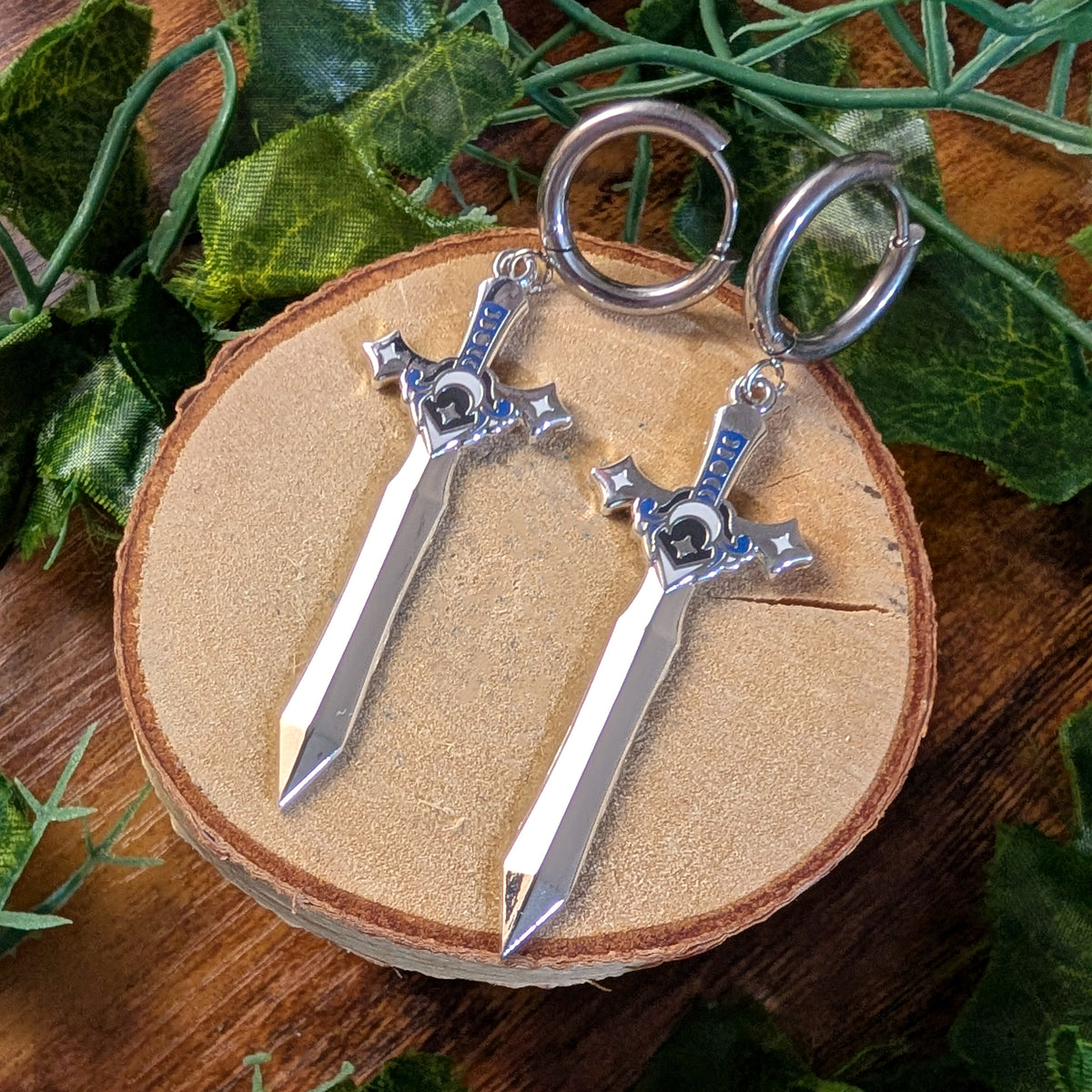 A pair of silver-colored keychains shaped like the Sword of Kings from The Legend of Zelda, displayed on a round wooden slice with green leaves in the background.