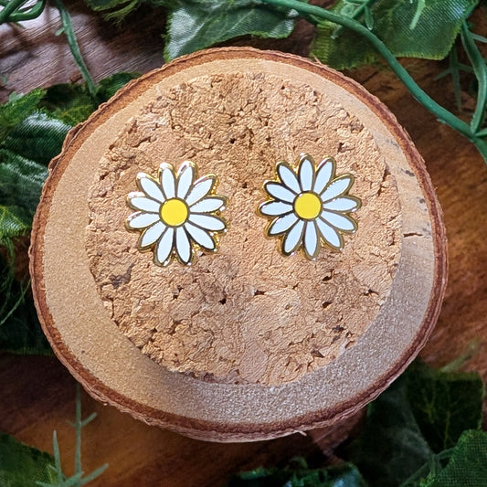 A pair of white daisy stud earrings with yellow centers, displayed on a round cork coaster surrounded by green leaves on a wooden surface.