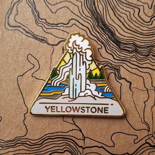 Triangle Yellowstone national park enamel pin featuring a view of Old Faithful geyser erupting with yellow sulfer hot springs in the background.