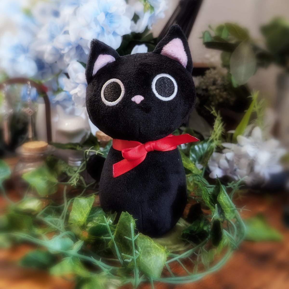 A black plush cat toy with pink inner ears, large white-rimmed eyes, a small pink nose, and a bright red bow around its neck, styled among green leaves and blurred blue flowers in the background.