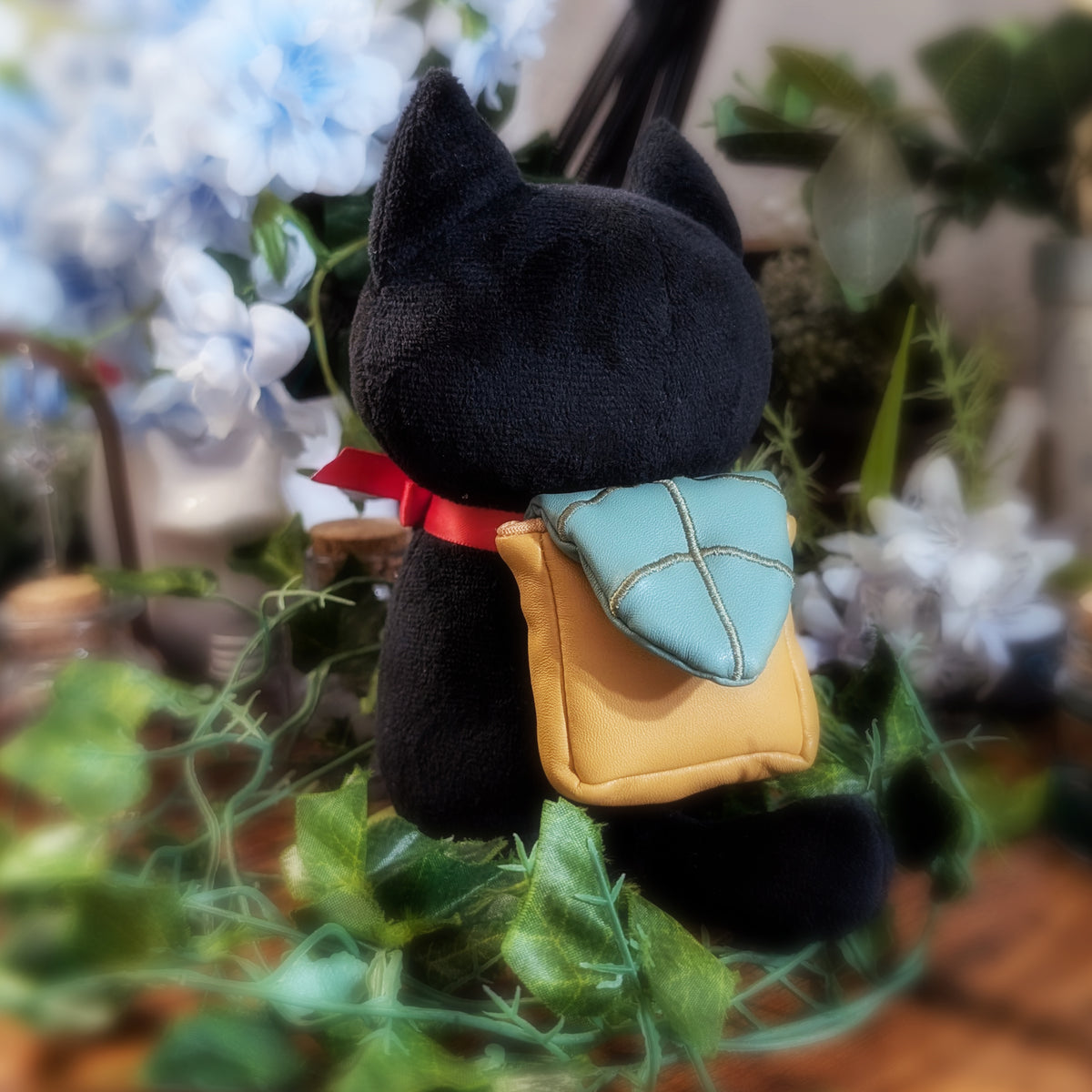 A black plush cat toy wearing a small yellow and teal backpack with a red ribbon, posed among green leaves and blurred blue flowers.