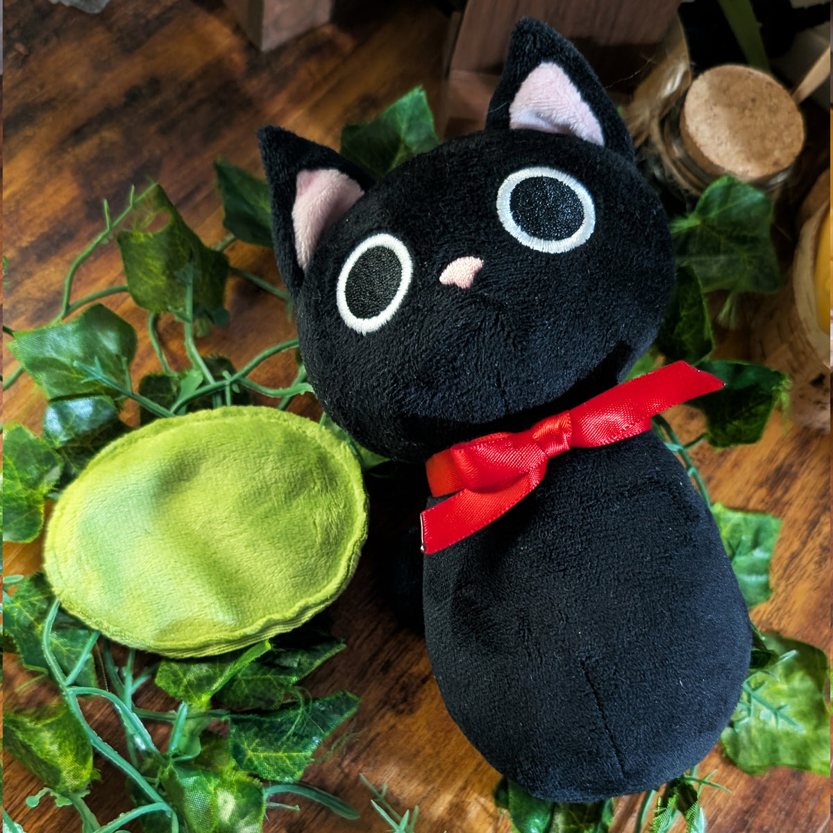 A black plush cat toy with embroidered white eyes, pink inner ears, and a red ribbon bow, displayed on a wooden surface with green ivy leaves and a green oval fabric prop.