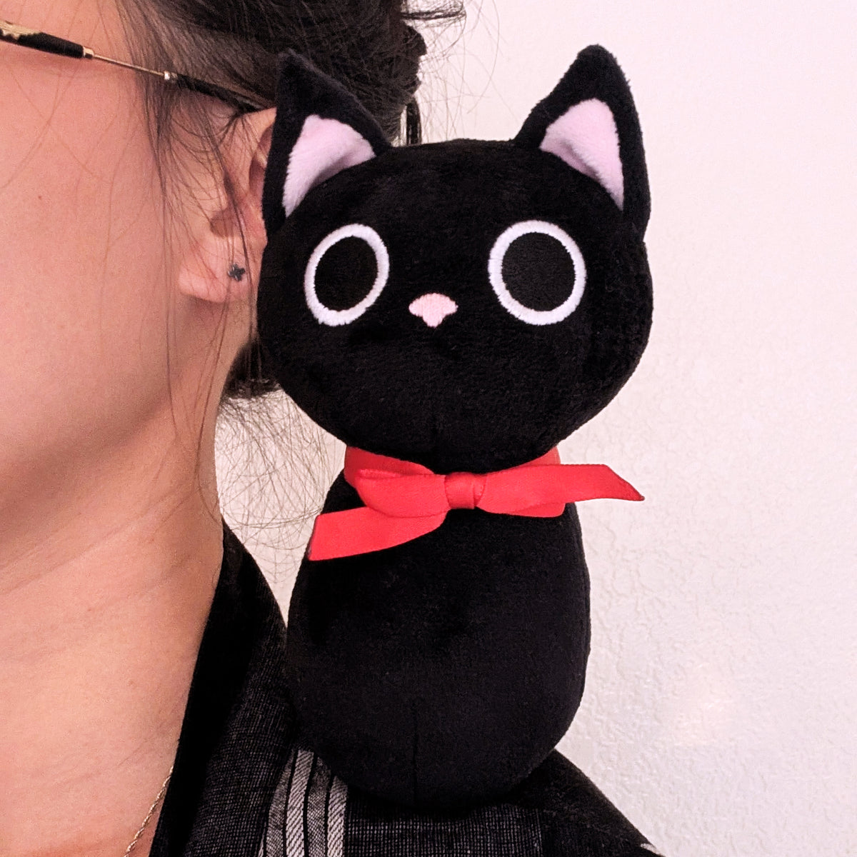 A black cat plush toy with pink inner ears, white-rimmed eyes, a small pink nose, and a red bow tie, shown sitting on a person's shoulder.