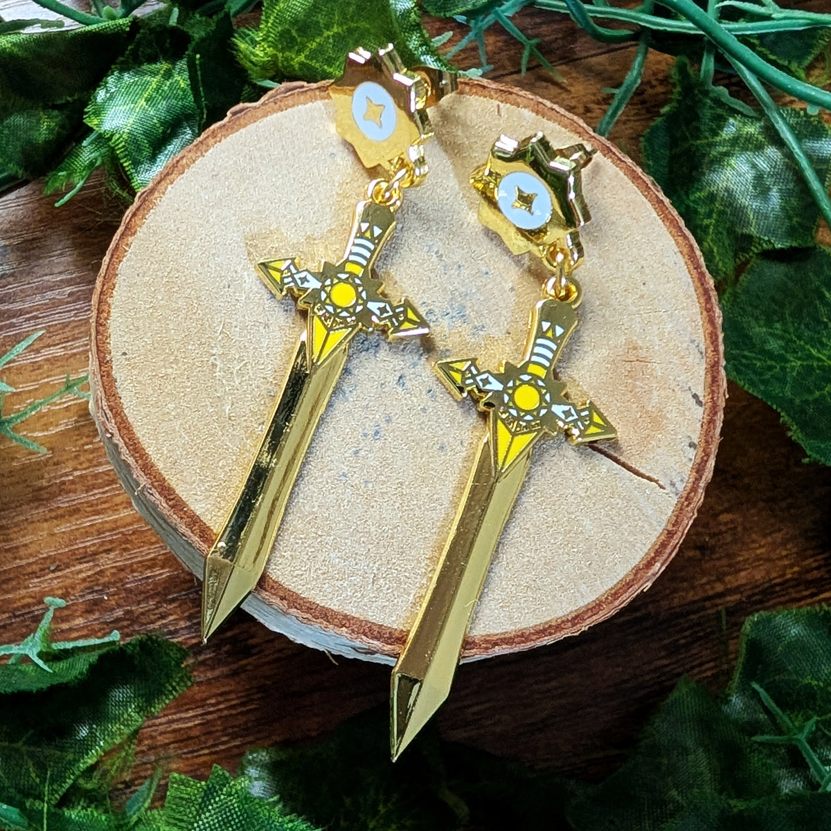 A pair of gold-toned dangle earrings shaped like miniature swords with shield pendants, displayed on a round wood slice surrounded by green leaves.