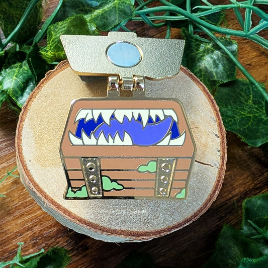 A small enamel pin shaped like an open treasure chest with a blue shark mouth inside, displayed on a round wood slice surrounded by green leaves on a wooden surface.