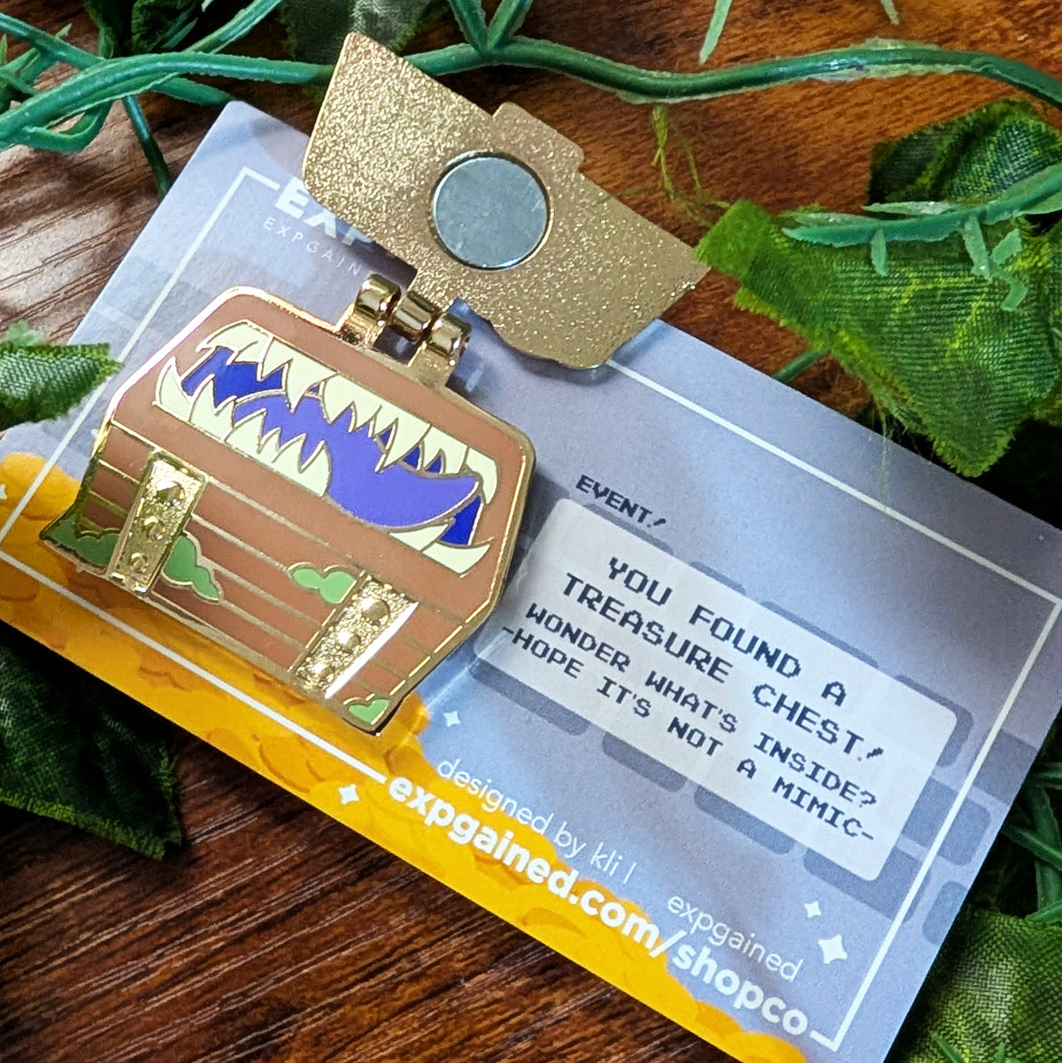 A treasure chest themed lapel pin with gold-tone metal, blue and green enamel details, attached to a branded display card. The pin is photographed on a wooden surface with decorative green leaves around it.