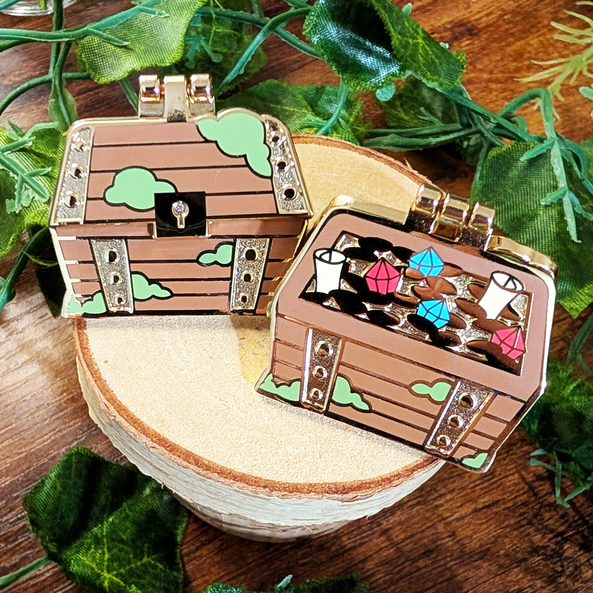Two miniature treasure chest enamel pins displayed on a wooden slice coaster with green foliage in the background. One pin is closed, showing a brown wood-grain texture, green clouds, and a gold clasp.