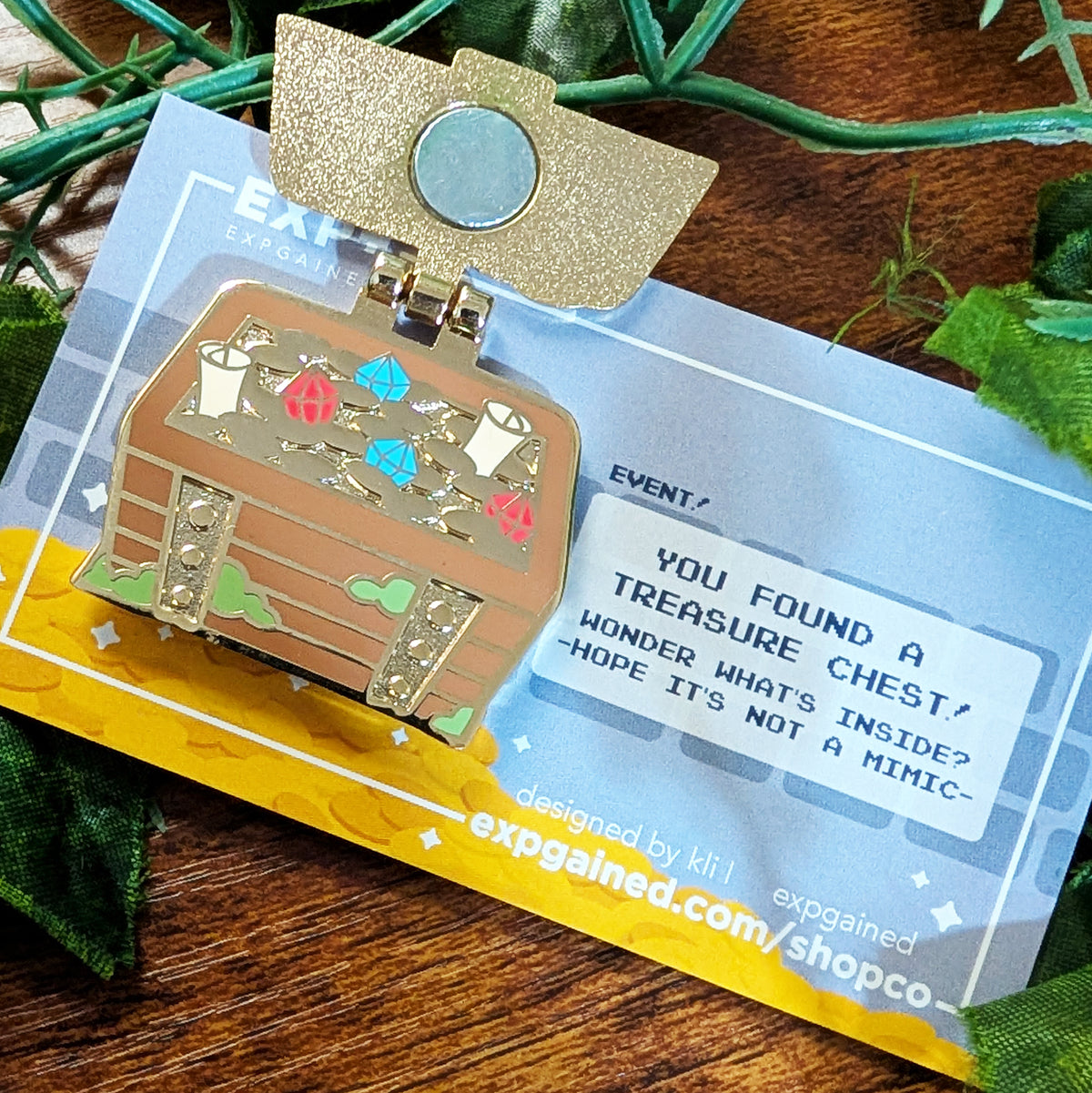 An enamel pin shaped like a treasure chest mounted on its original display card, photographed on a wooden surface with green leaves around it. The pin is gold-toned with multicolor accents including blue, red, and green, and the card has text that reads 'YOU FOUND A TREASURE CHEST! WONDER WHAT'S INSIDE? HOPE IT'S NOT A MIMIC'.