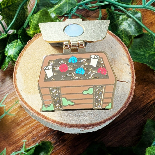 A small illustrated treasure chest pin with an open lid sits on a round wood slice. The chest has gold-toned trim, brown wood planks, colorful gem accents in blue, pink, and red, and green cloud details.